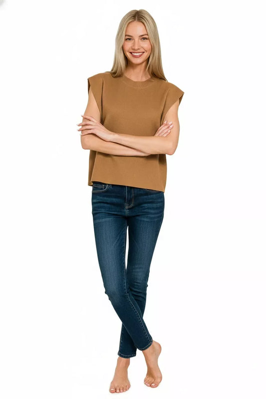 Woman in sleeveless camel knit top and dark skinny jeans standing against white background