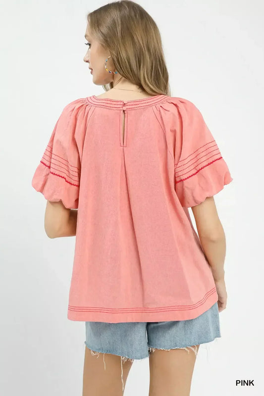 Back view of woman in a pink puff sleeve blouse with red stitching, paired with denim shorts