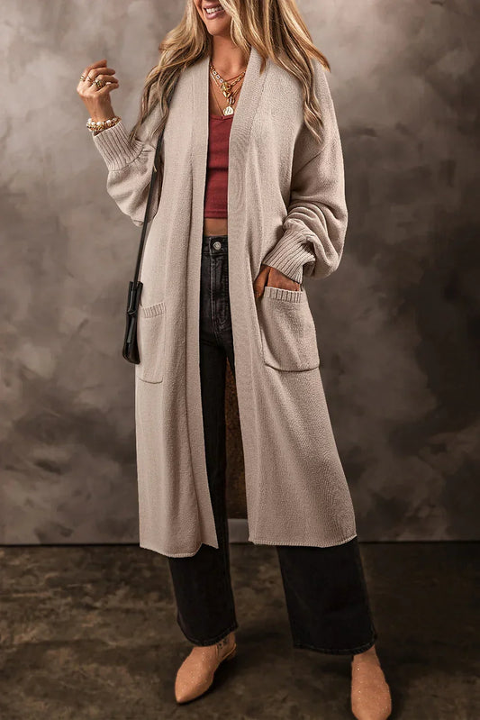 Woman wearing a long beige knit cardigan with pockets, dark jeans, and tan pointed shoes