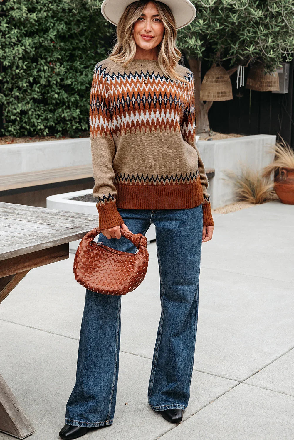 Woman in geometric patterned sweater, wide-leg jeans, and hat, holding a woven brown handbag outdoors.