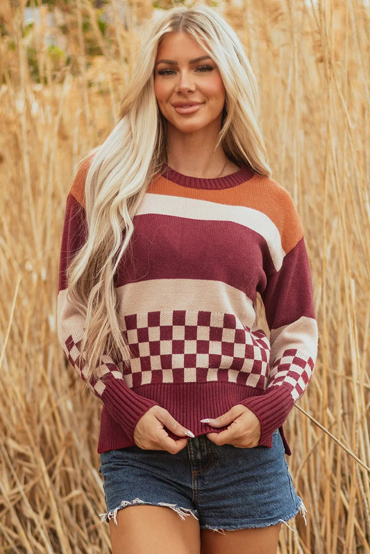 Woman wearing a burgundy, beige, and orange striped and checkered knit sweater with denim shorts, standing outdoors in tall dry grass.