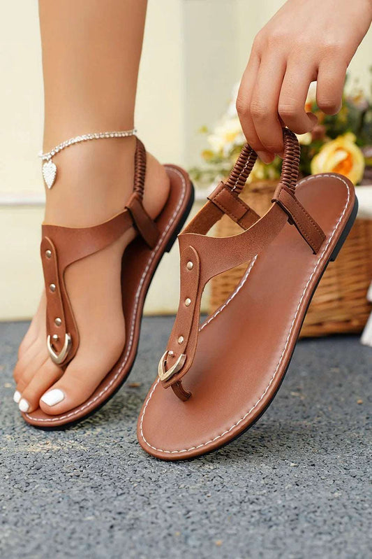 Brown flat thong sandals with gold accents, ankle strap, and stitched sole, worn outdoors.
