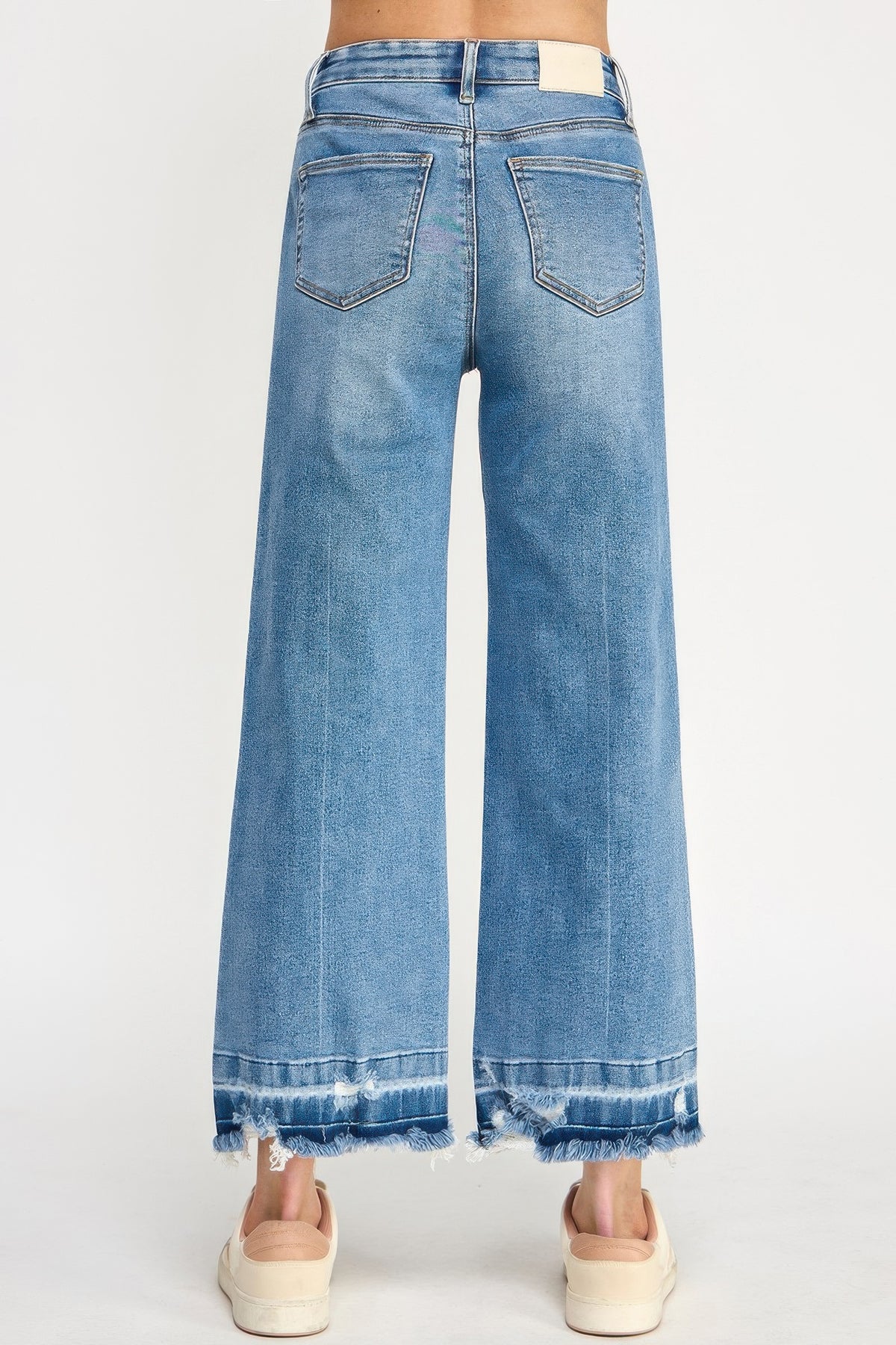 A woman wearing Tummy Control Cropped Wide Leg Jeans with a frayed hem and white sneakers against a white background.