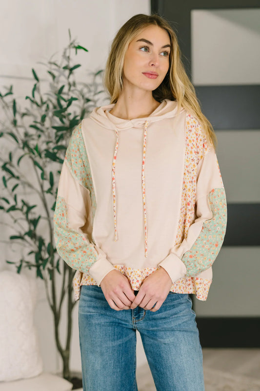 Woman wearing a floral patchwork hooded pullover with pastel sleeves and blue jeans.