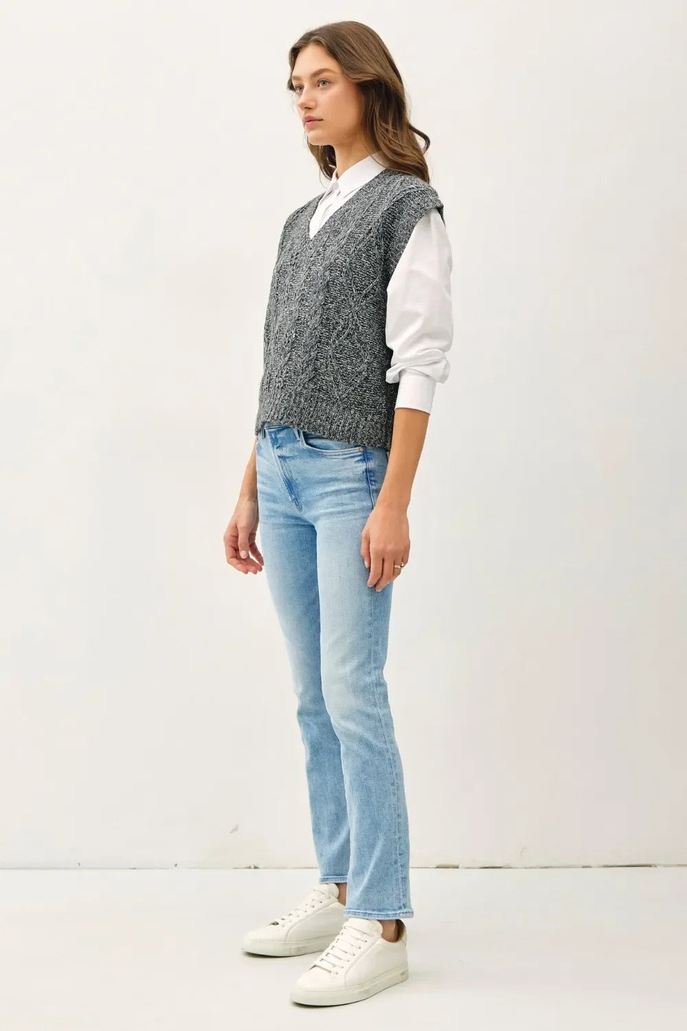 Woman wearing a gray knit vest over a white shirt, light blue jeans, and white sneakers