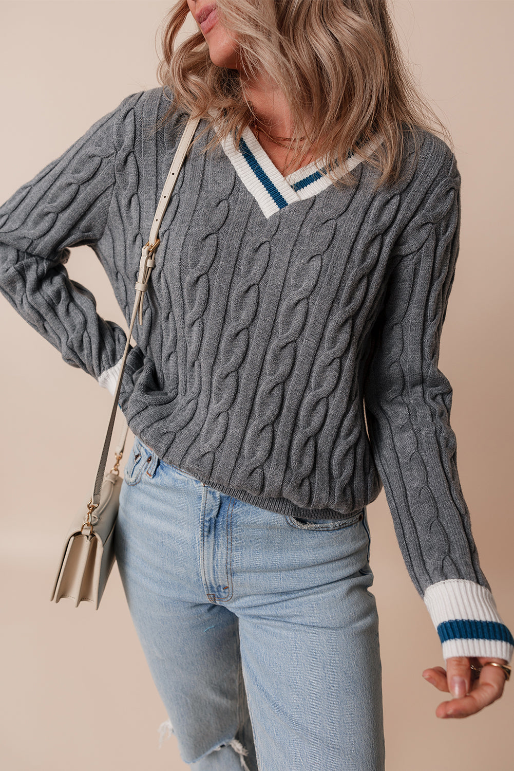 Person wearing a gray cable knit sweater with blue jeans and a beige background