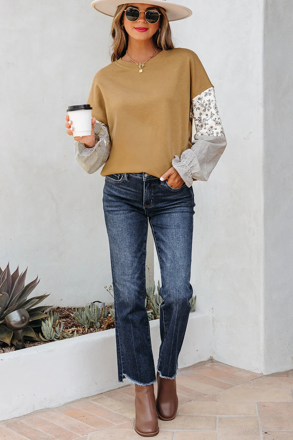 Woman in a tan long sleeve top with floral and striped sleeves, dark jeans, brown ankle boots, wide hat, and sunglasses, holding a coffee cup outdoors.