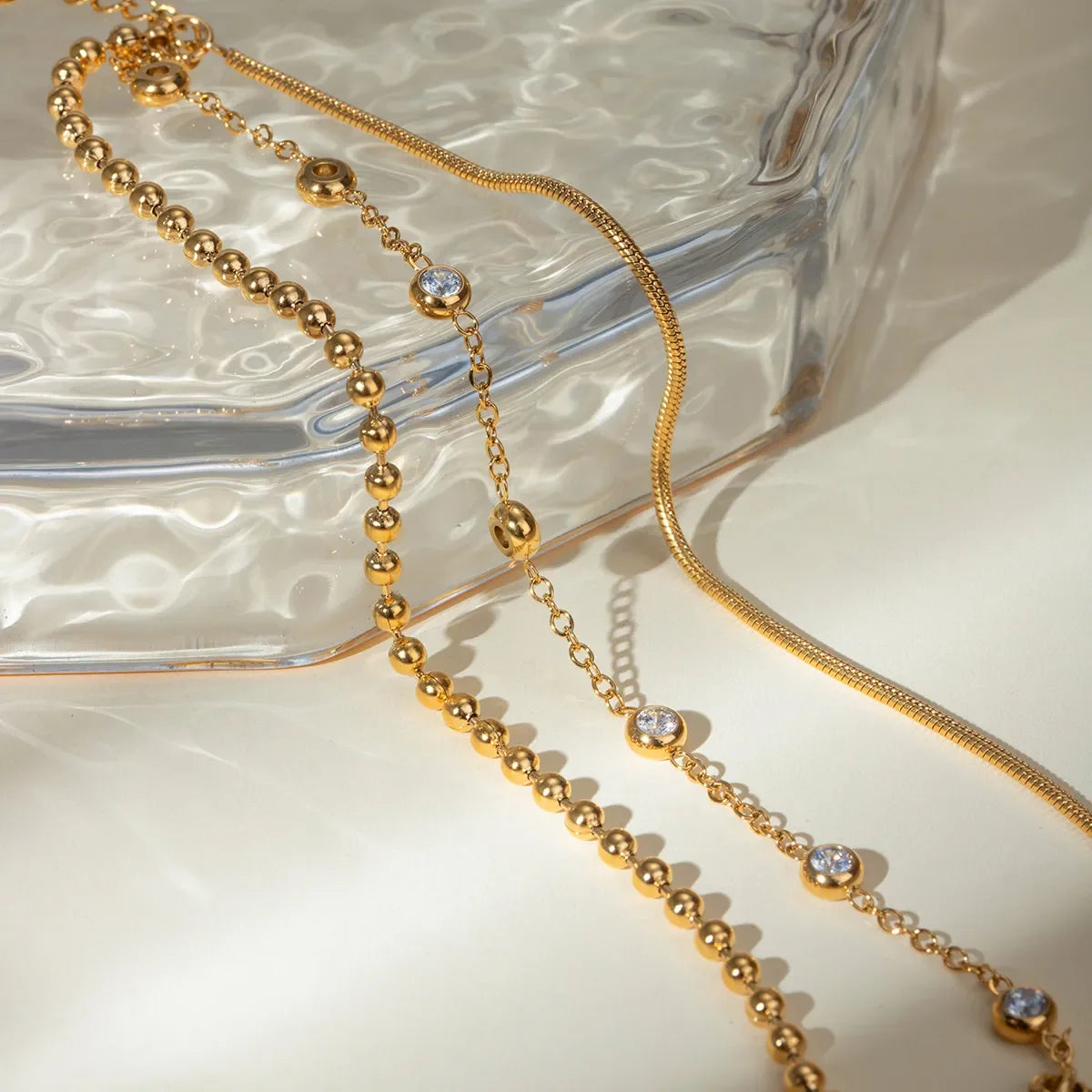 Gold chain necklaces with beaded and crystal accents displayed on a textured glass tray.