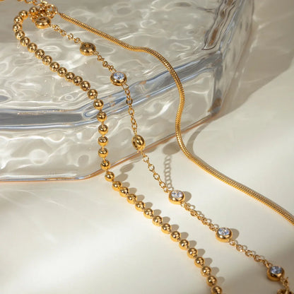 Gold chain necklaces with beaded and crystal accents displayed on a textured glass tray.