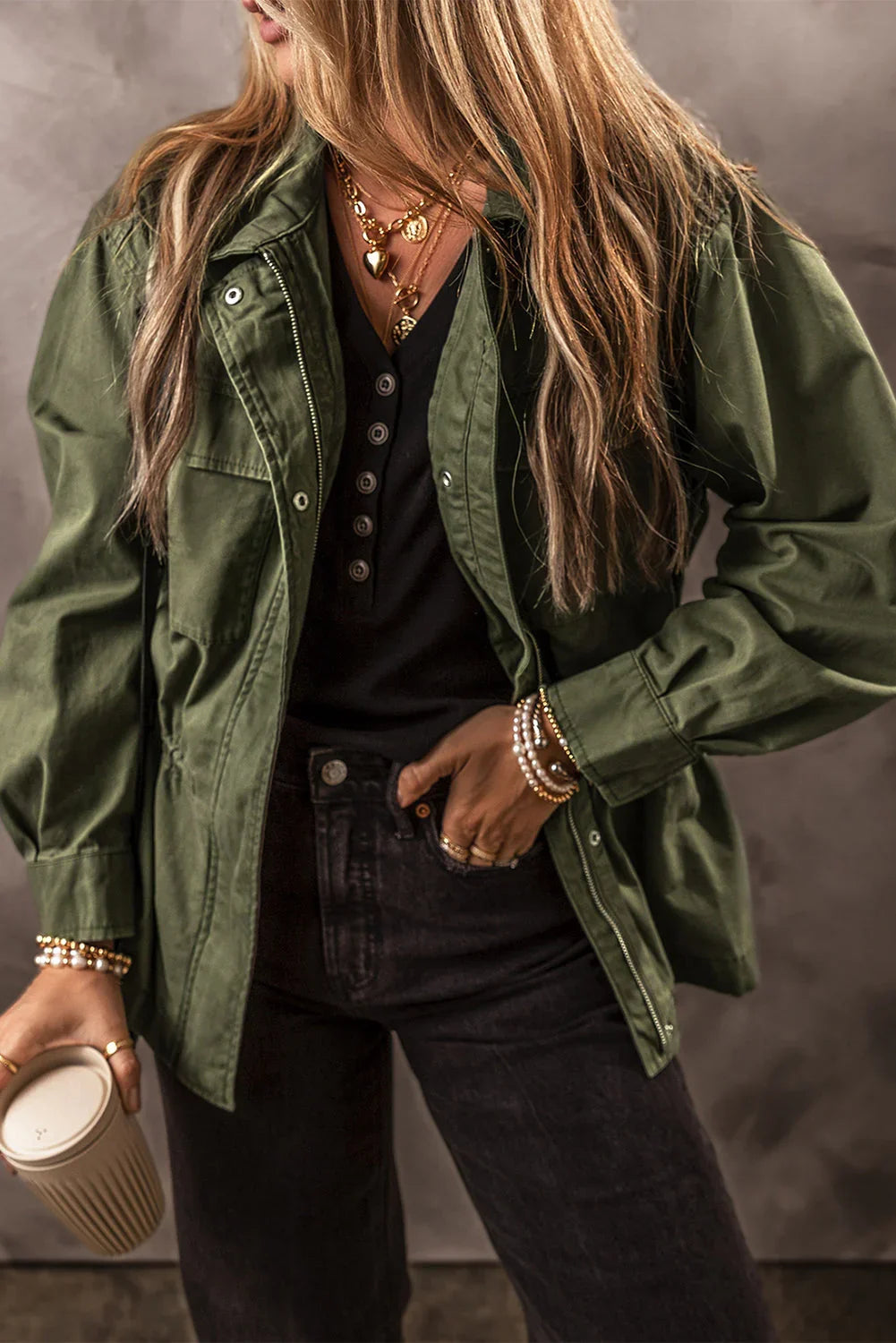 Woman in an olive green jacket, black top, and jeans with layered gold jewelry