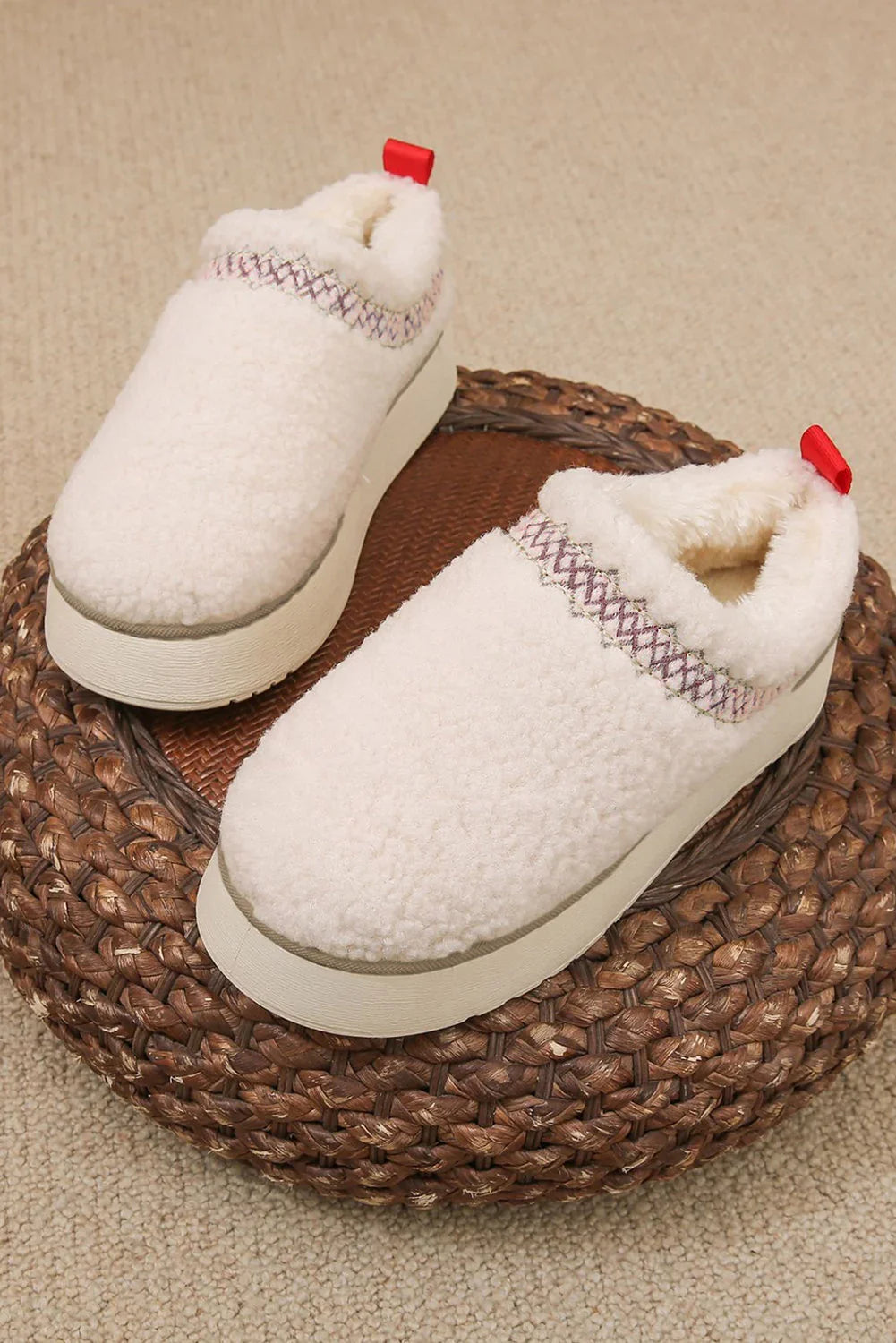 Cream sherpa slip-on platform slippers with red pull tabs and decorative stitching, on woven pouf