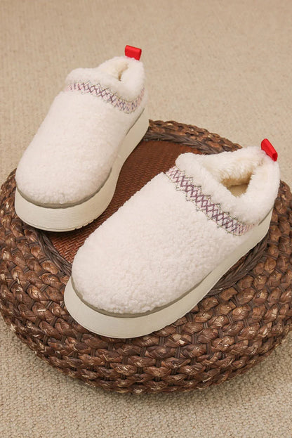 Cream sherpa slip-on platform slippers with red pull tabs and decorative stitching, on woven pouf