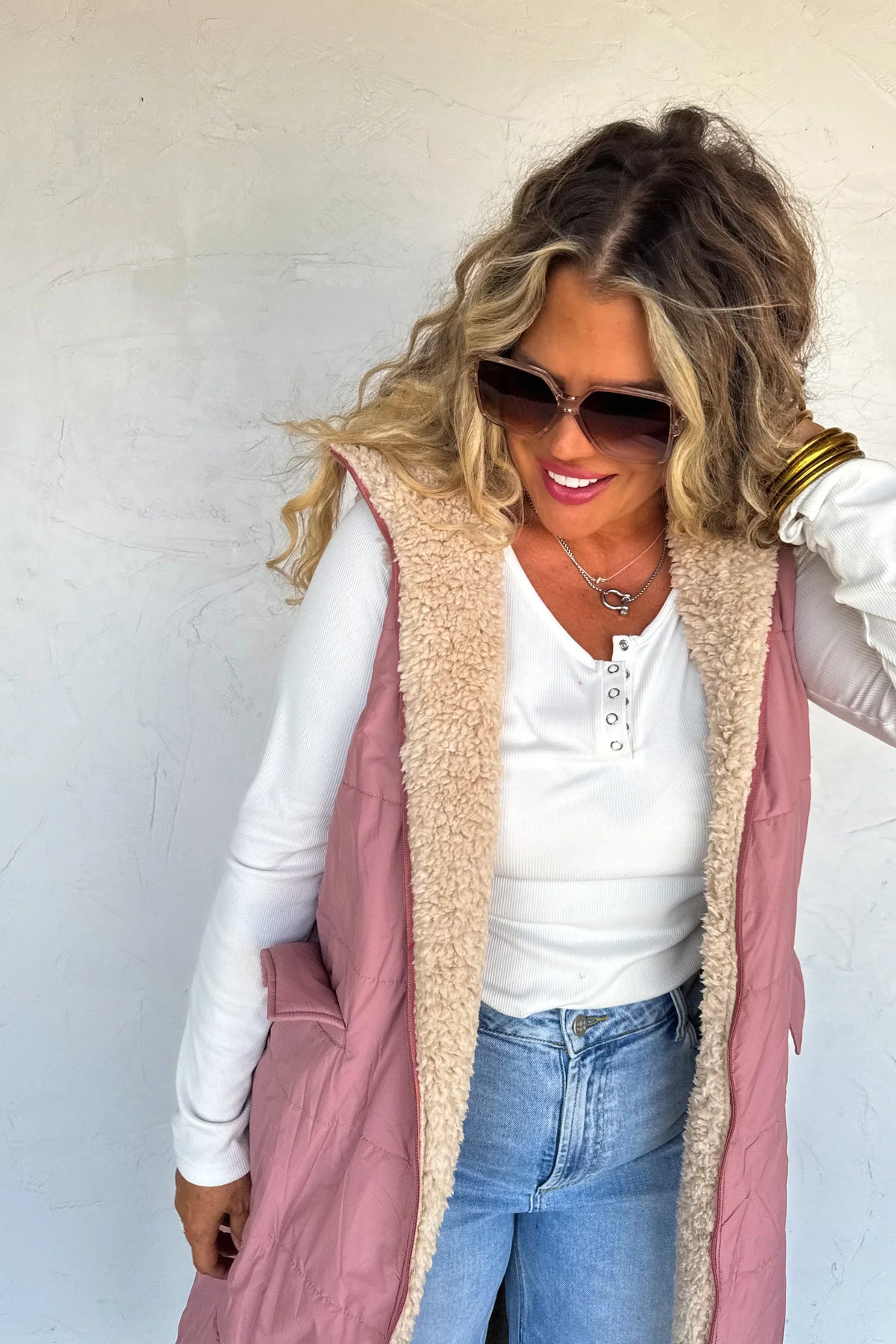 Woman in a pink sherpa-lined vest, white long-sleeve top, and blue jeans, wearing sunglasses.