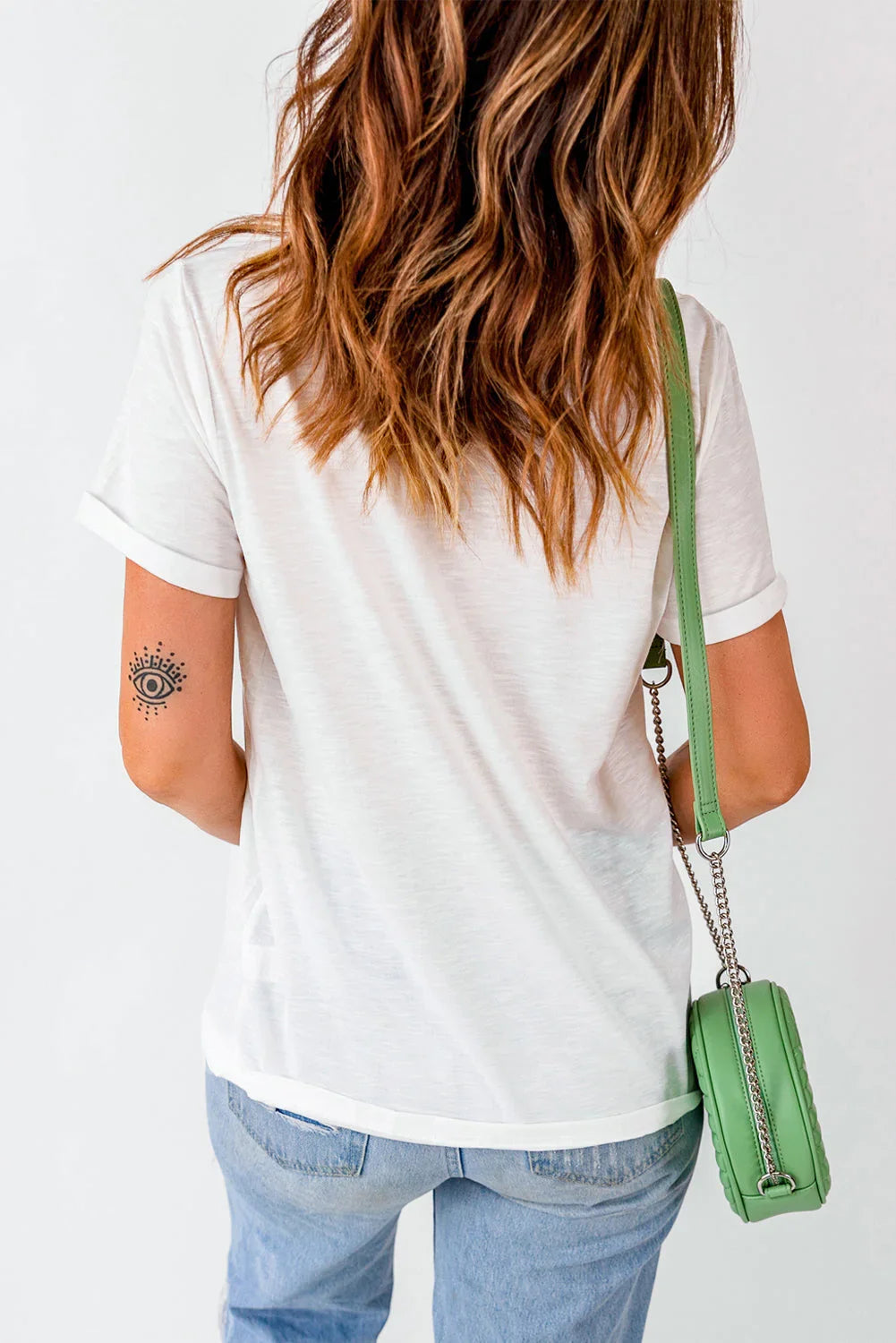 Woman in white t-shirt and light blue jeans, green crossbody bag, wavy hair, arm tattoo