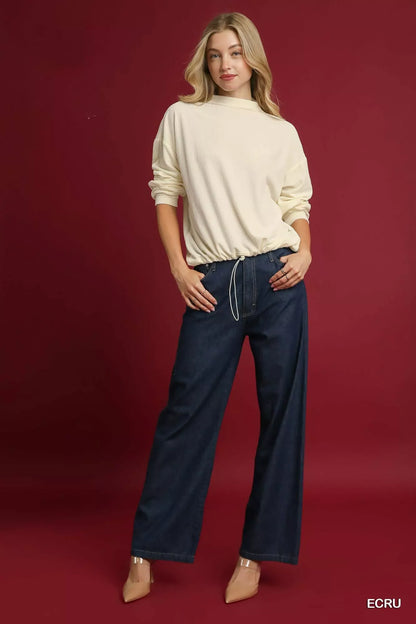 Woman in relaxed cream sweater and wide-leg dark denim pants, standing against a burgundy background.