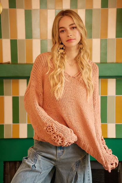Woman in peach knit sweater with floral cuffs and relaxed jeans, standing indoors by colorful tiled wall