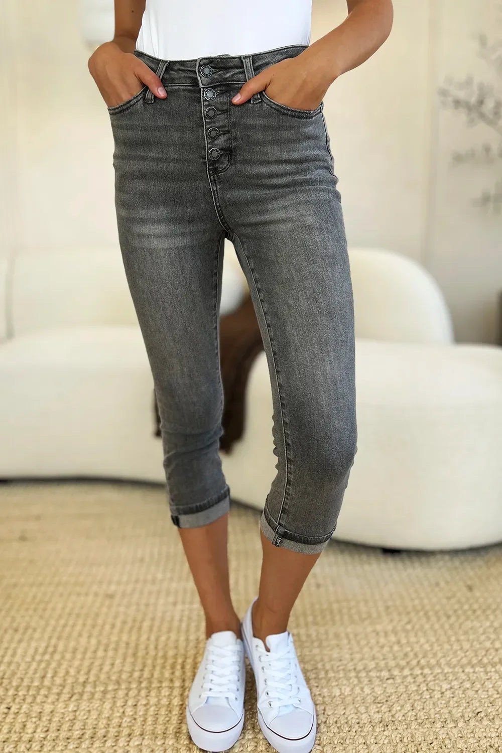 Woman wearing grey cropped button-fly jeans and white sneakers in a modern living room.