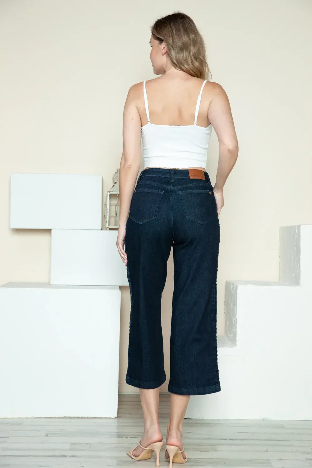 Woman in a white camisole and dark wide-leg cropped jeans standing indoors, back view