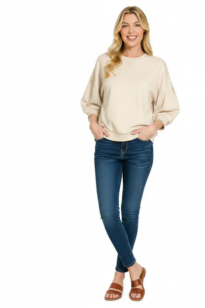 Woman wearing a beige pullover sweater, fitted blue jeans, and tan sandals on white background.