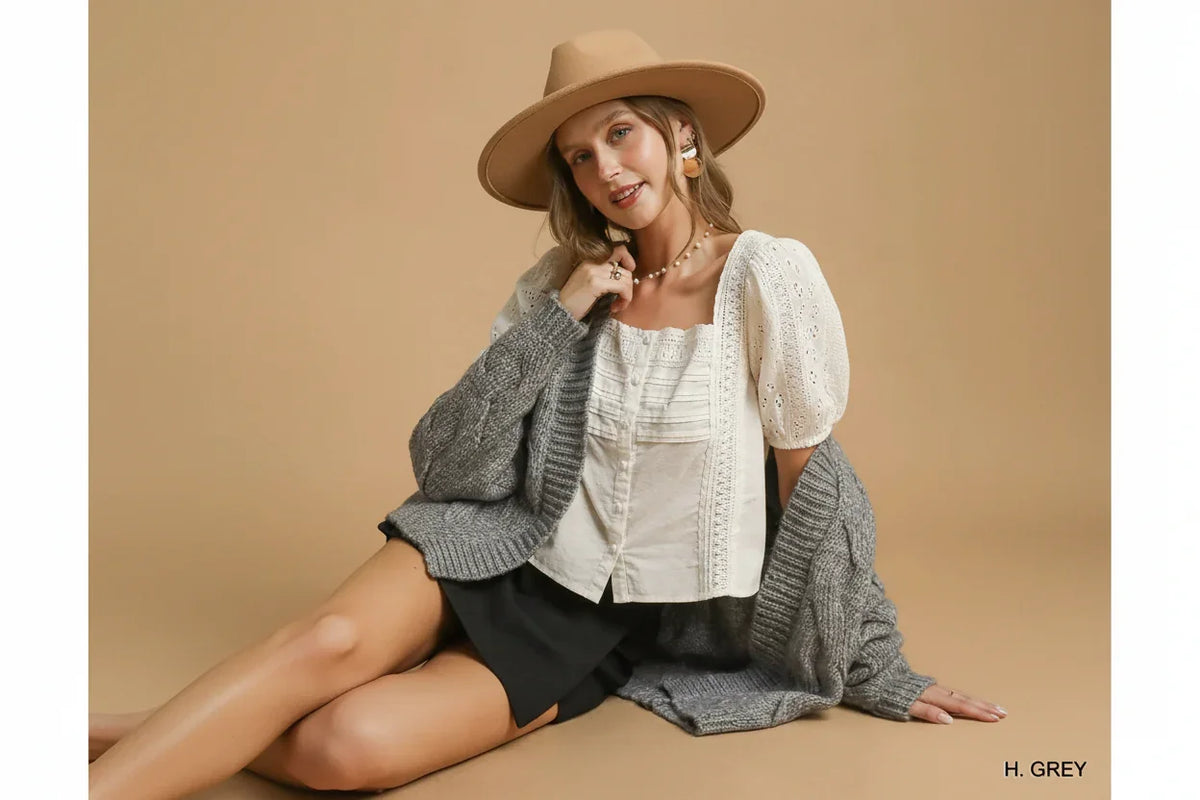 Woman in neutral hat, white eyelet blouse, grey knit cardigan, and black shorts sitting on tan backdrop
