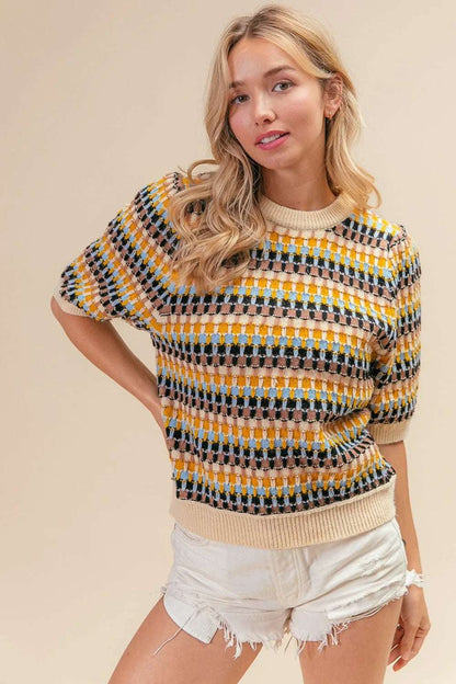 Woman wearing a patterned short sleeve knit sweater with beige, yellow, blue, and black checks, paired with white denim shorts.