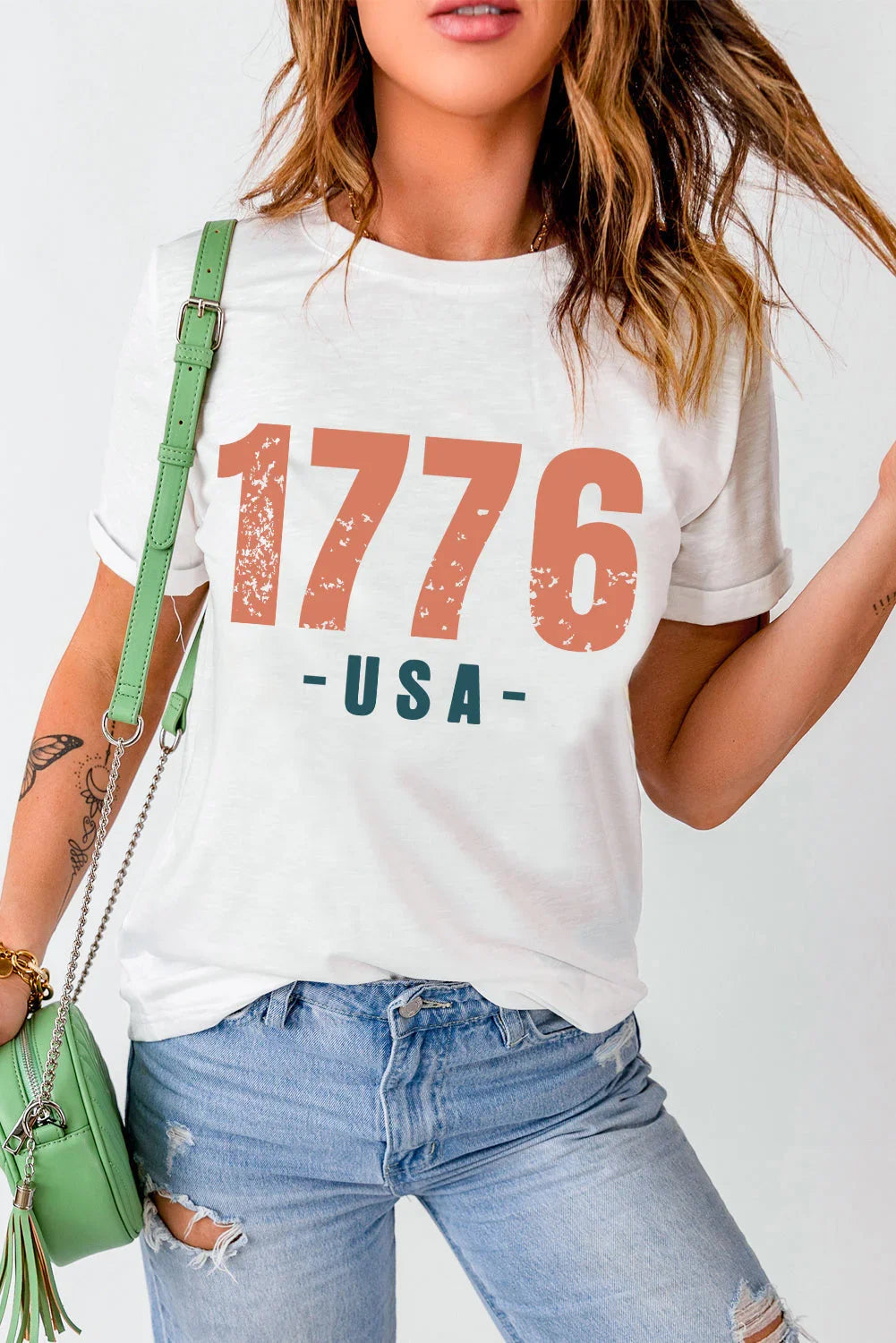 Woman wearing white 1776 USA graphic t-shirt with ripped light denim jeans and green purse