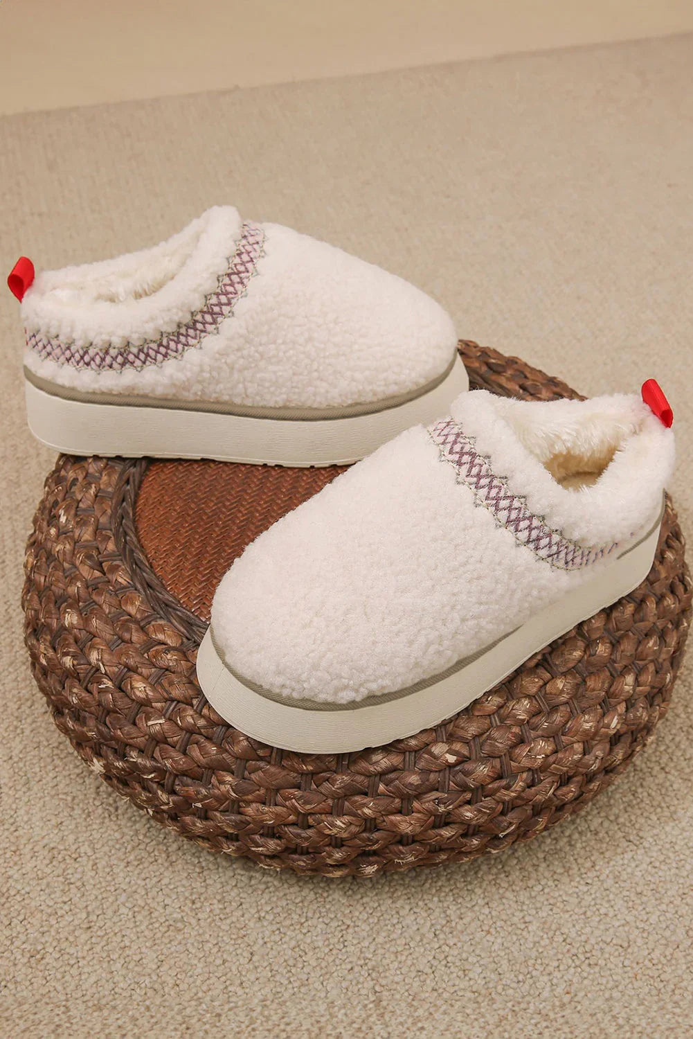 Pair of cozy white sherpa slip-on slippers with embroidered trim and red pull tabs on a woven ottoman