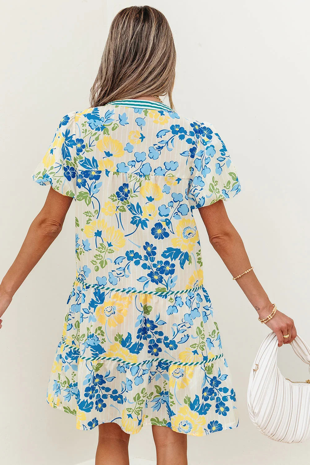 Woman in a short-sleeve floral dress with blue, yellow, and green flowers, holding a white purse.