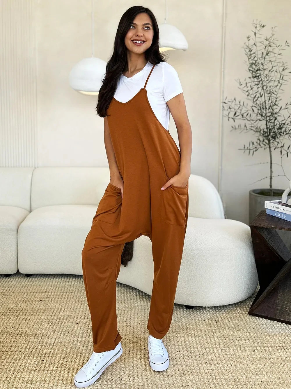 Woman wearing rust brown relaxed jumpsuit over white t-shirt and white sneakers in a modern living room