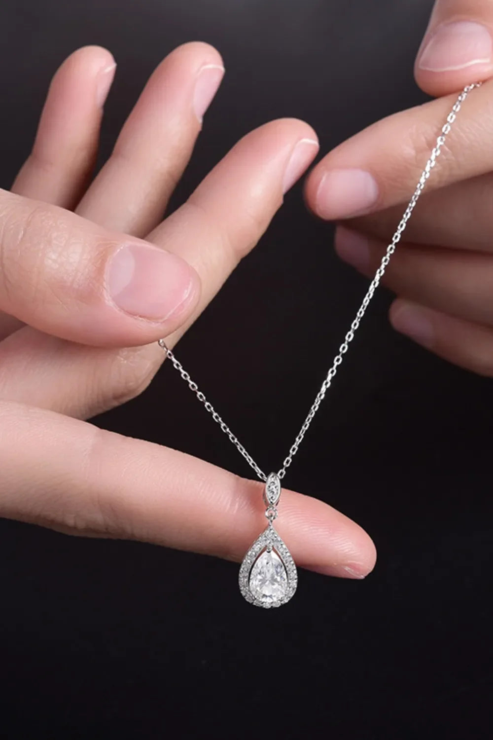Hand holding a silver chain necklace with teardrop-shaped crystal pendant on black background