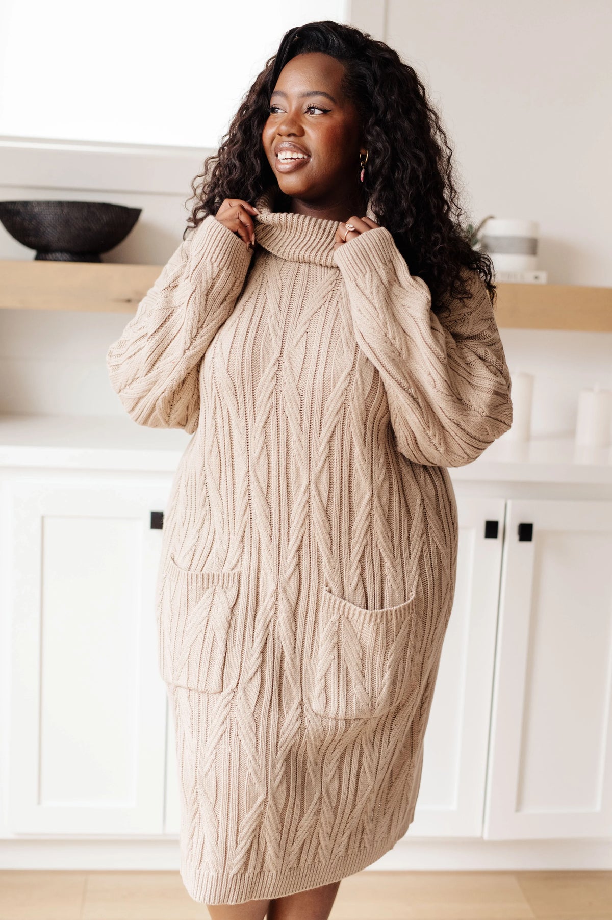 Woman modeling a beige cable knit sweater dress with pockets in a modern kitchen.