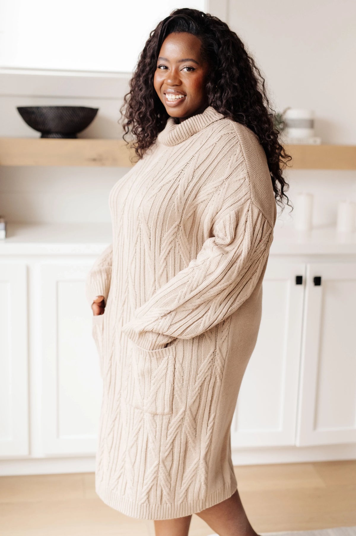 Woman wearing a beige cable knit sweater dress in a bright, modern kitchen