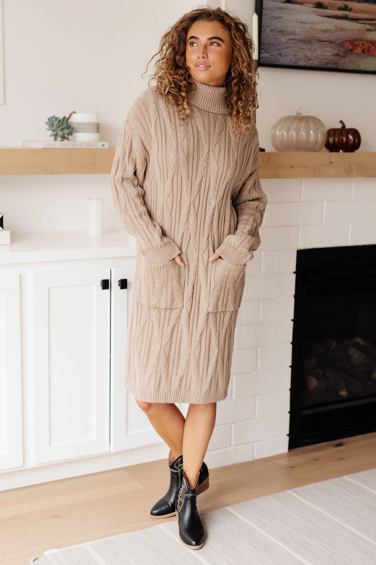Woman wearing a beige cable knit sweater dress with pockets and black ankle boots indoors