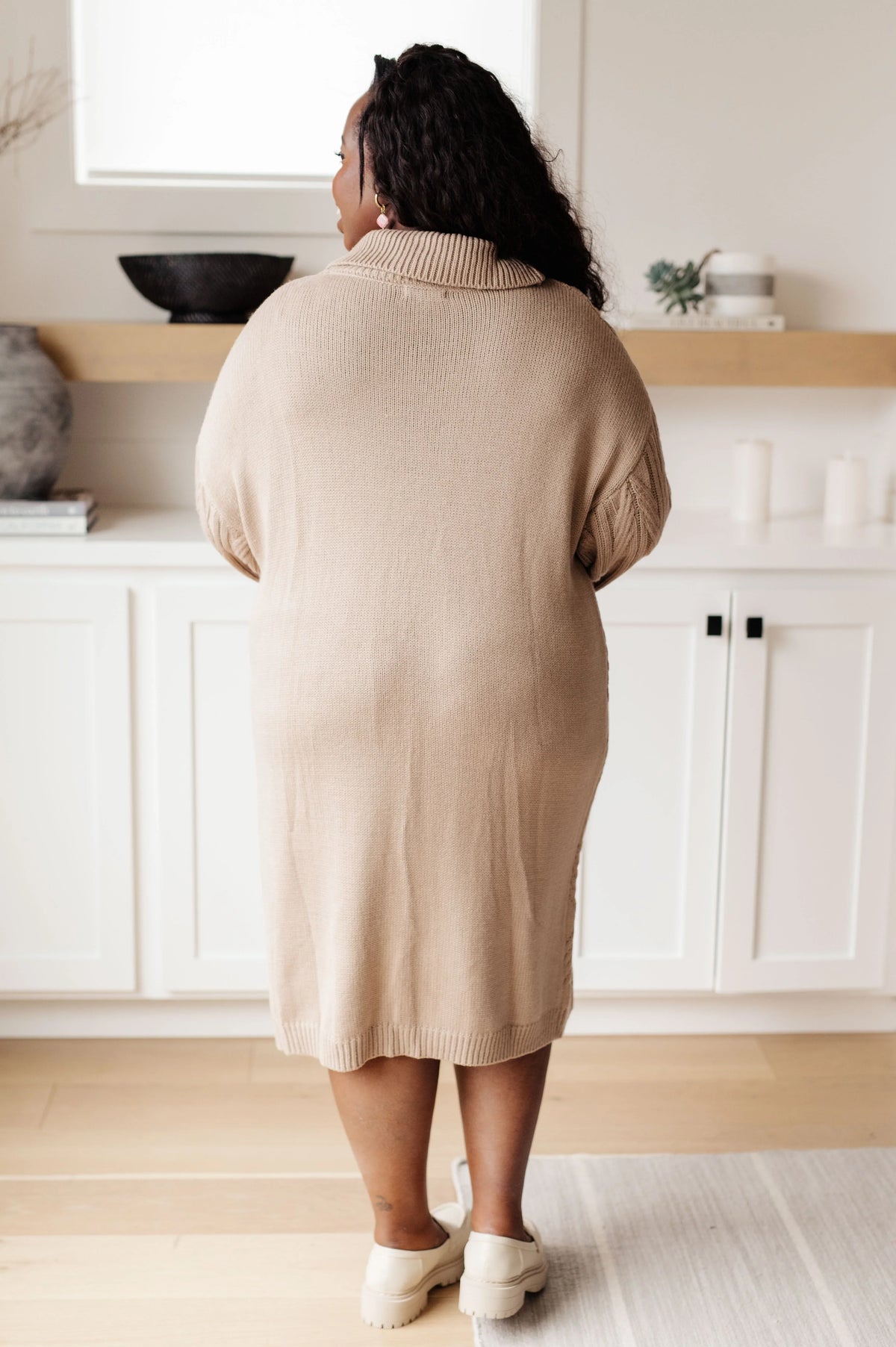 Woman wearing a beige knit sweater dress with long sleeves, shown from the back in a modern living space.