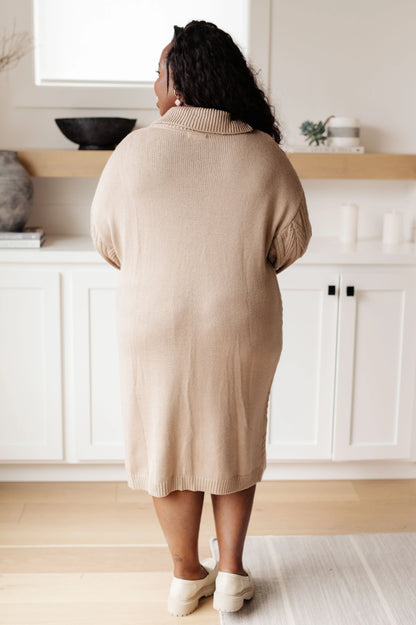 Woman wearing a beige knit sweater dress with long sleeves, shown from the back in a modern living space.