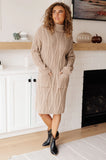 Woman wearing a beige cable knit sweater dress with pockets and black ankle boots indoors