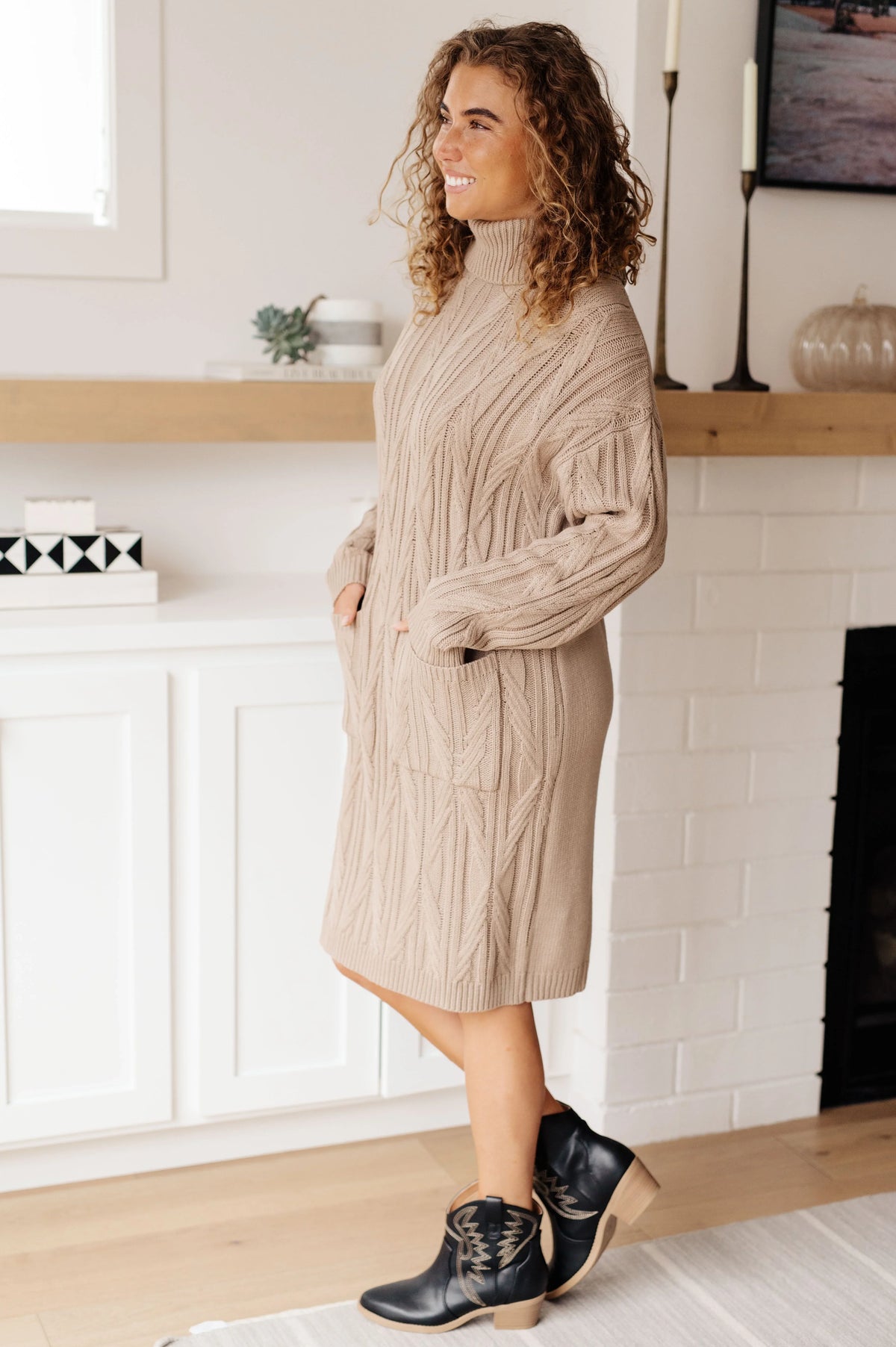 Woman wearing a beige cable knit sweater dress with black ankle boots in a modern living room.