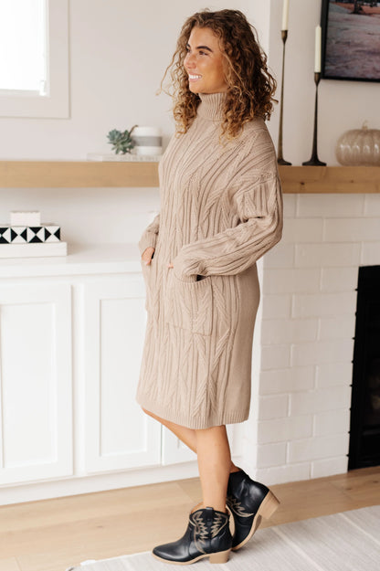 Woman wearing a beige cable knit sweater dress with black ankle boots in a modern living room.