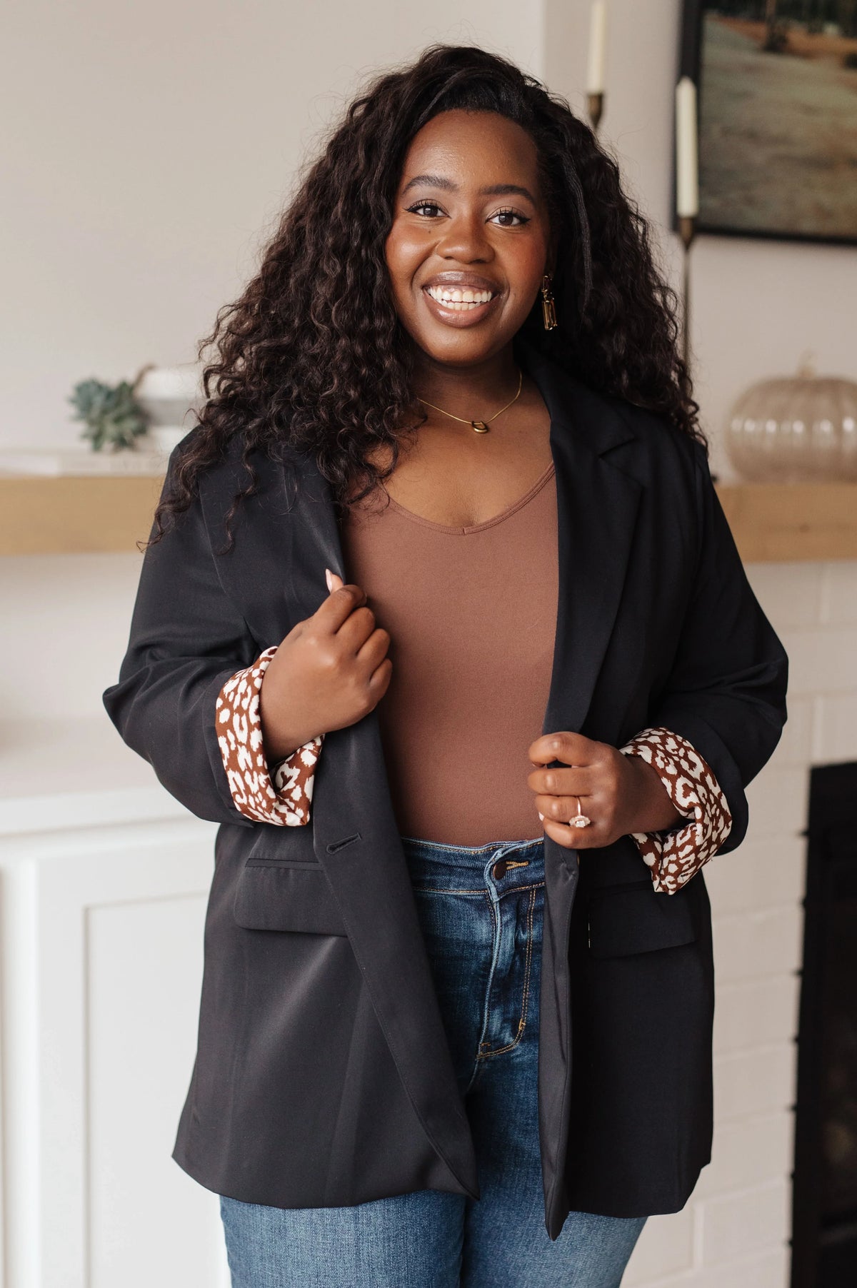 Woman in black blazer with patterned cuffs, brown top, and jeans, smiling in a modern living room