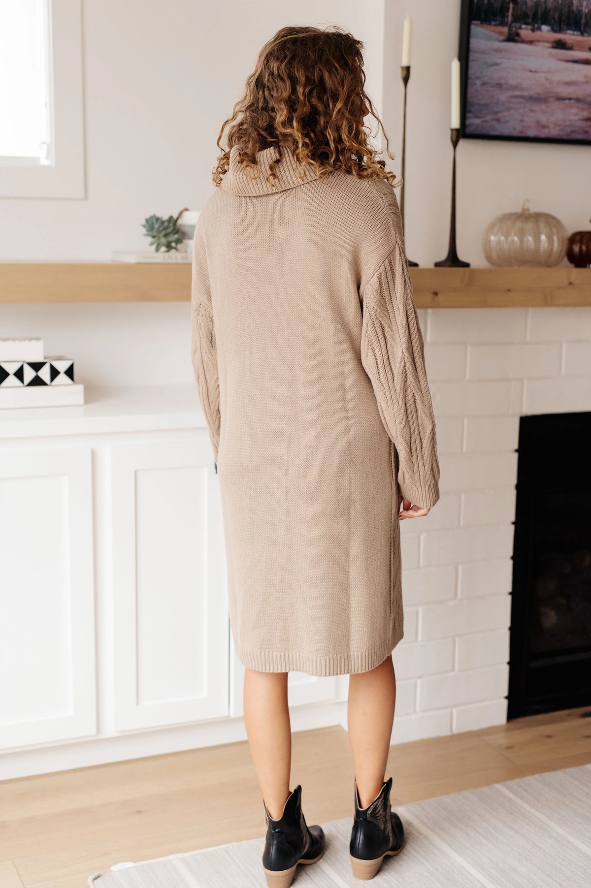Woman in a beige knit sweater dress with cable sleeves standing in a modern living room