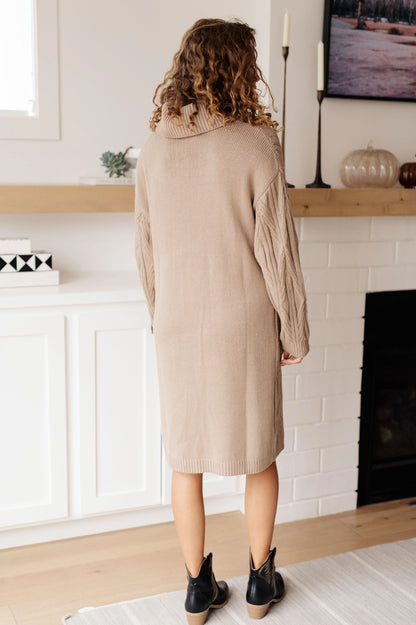 Woman in a beige knit sweater dress with cable sleeves standing in a modern living room
