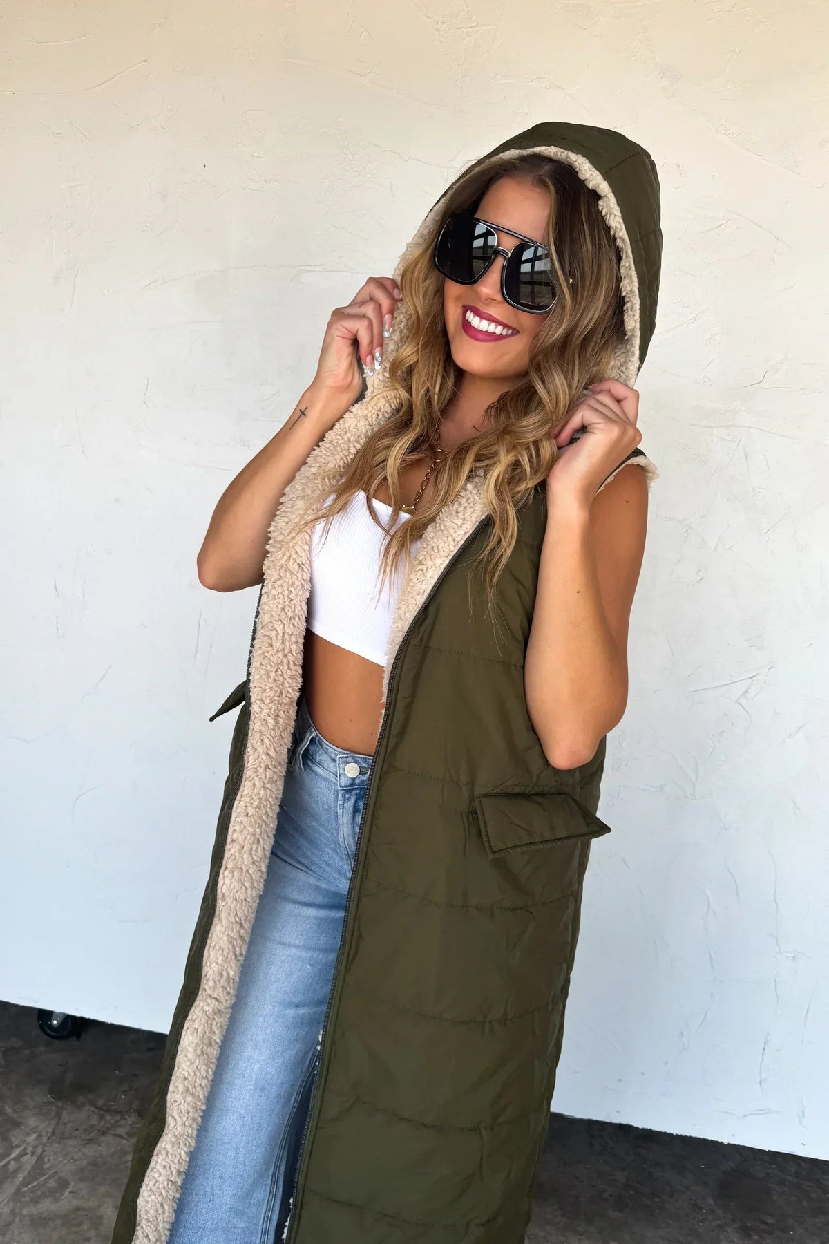 Woman wearing olive green hooded sherpa-lined vest, white crop top, and blue jeans