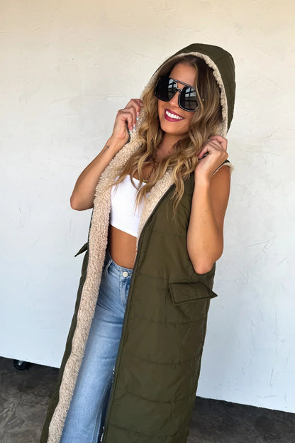 Woman wearing olive green hooded sherpa-lined vest, white crop top, and blue jeans