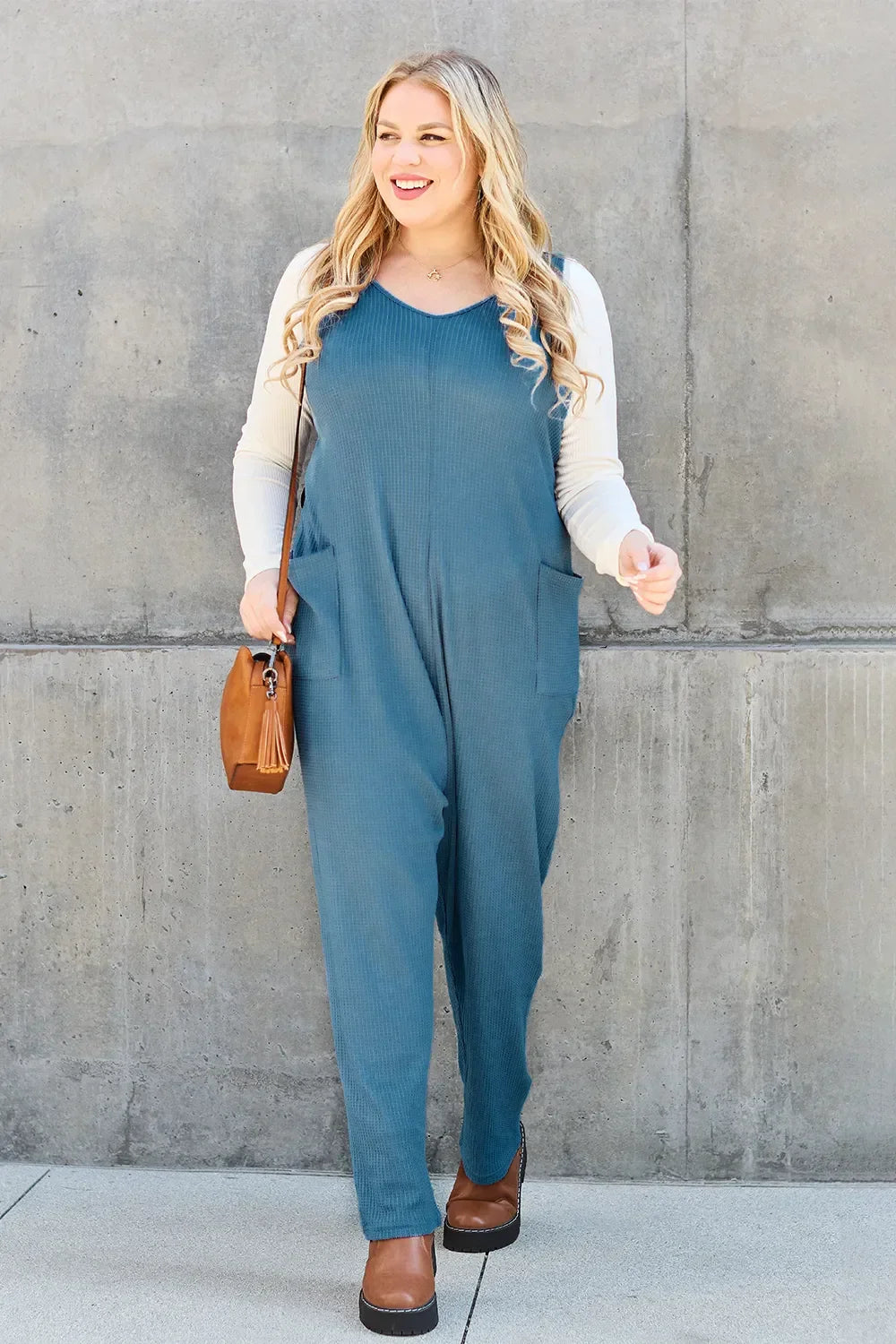Woman in blue sleeveless jumpsuit with cream top, brown purse and shoes, standing by concrete wall
