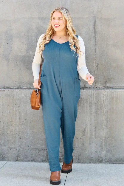 Woman in blue sleeveless jumpsuit with cream top, brown purse and shoes, standing by concrete wall