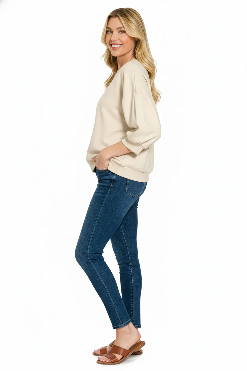 Woman wearing a cream pullover top, blue skinny jeans, and brown sandals against white background