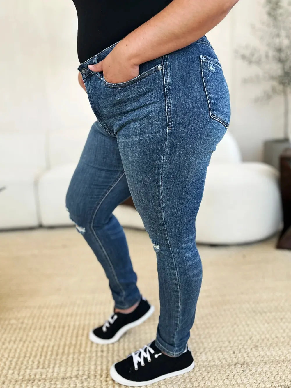 Woman wearing mid-rise skinny blue jeans with slight distressing and black sneakers indoors