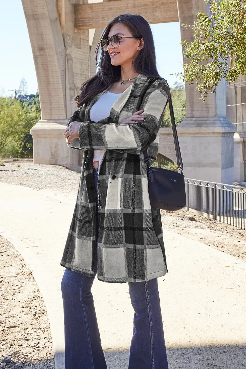Woman outdoors wearing black and white plaid coat, flared jeans, sunglasses, and crossbody bag