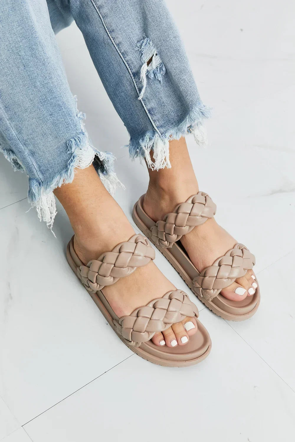 Woman wearing beige double-strap braided sandals and distressed light wash jeans on tile floor.