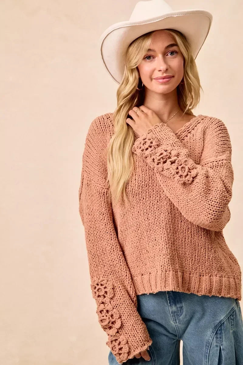 Woman wearing a blush pink chunky knit sweater with floral sleeve details and a white hat